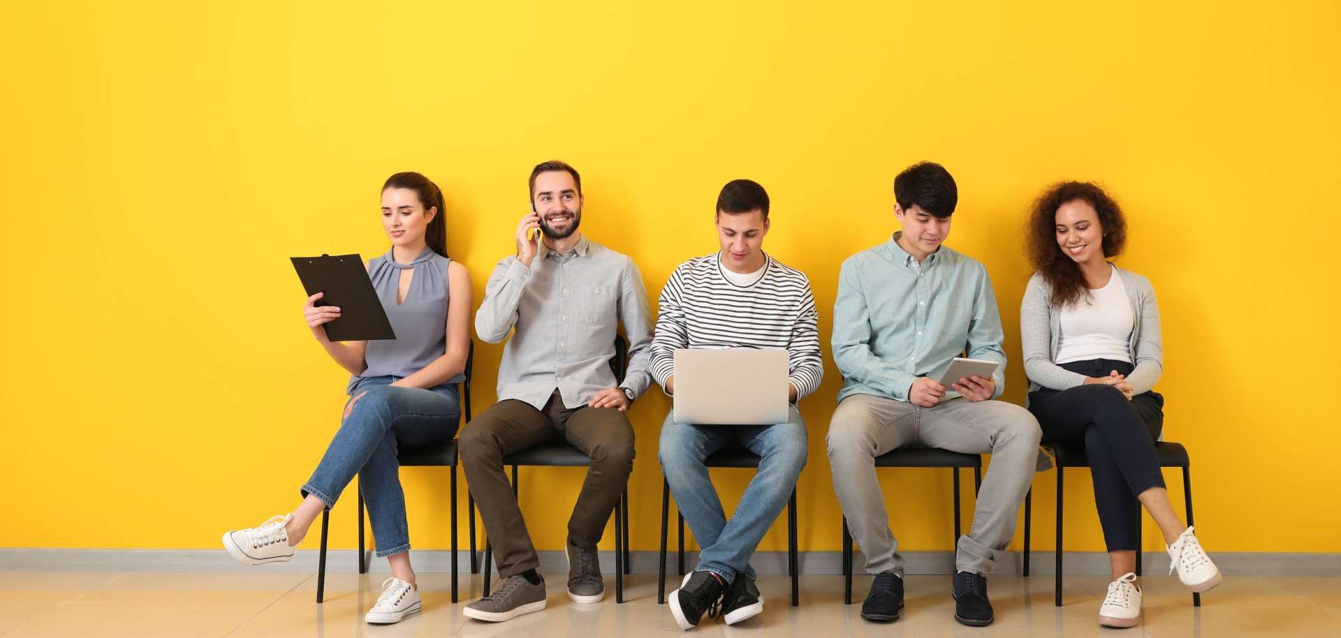 Das Bild zeigt junge Menschen mit mobilen Geräten wie einem Laptop oder einem Handy, die vor einer gelben Wand auf ein Vorstellungsgespräch warten.