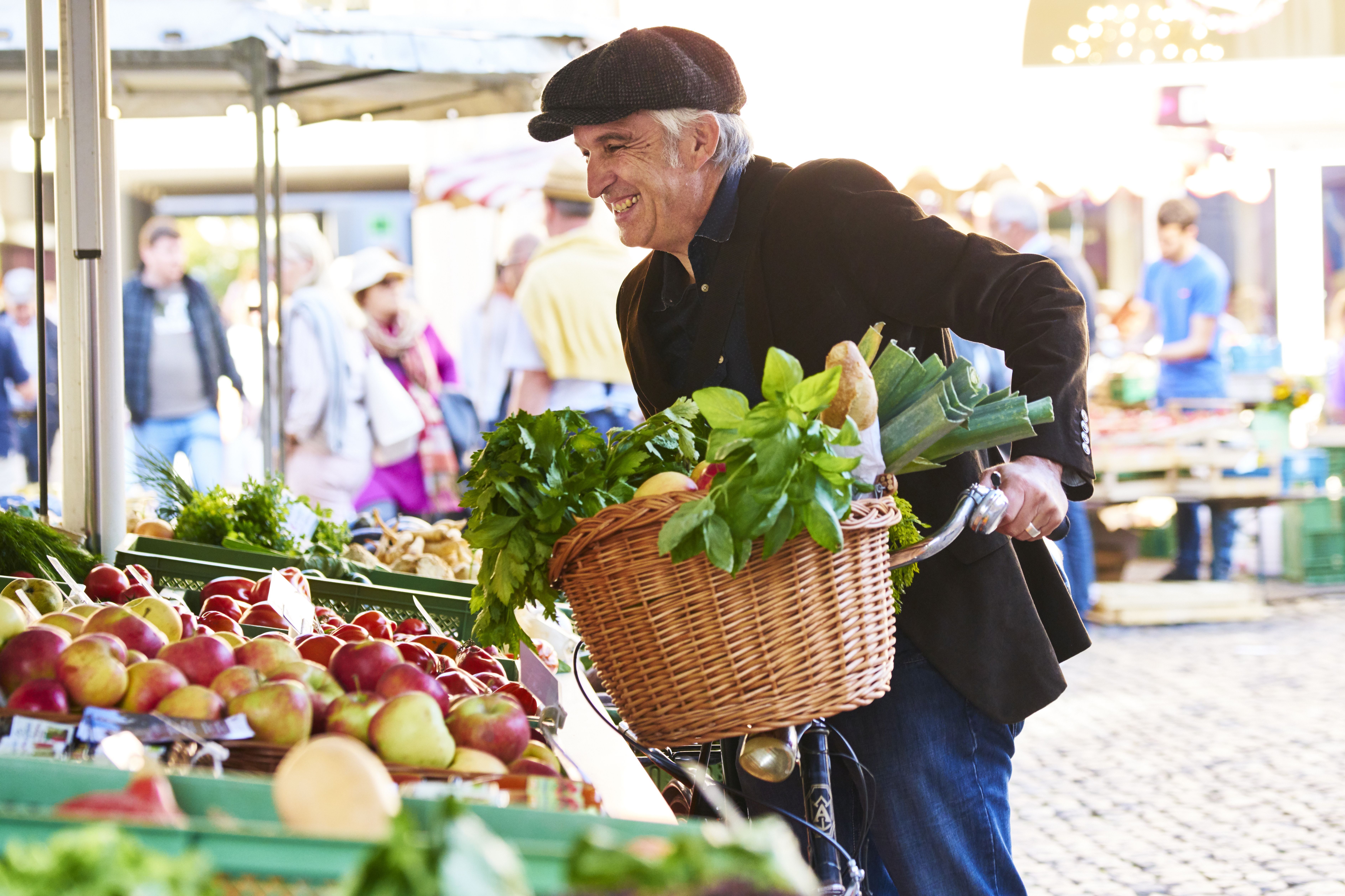 Ein Mann mit Fahrrad auf dem Markt in einer Stadt mit seinen Einkäufen im Fahrradkorb, darunter Baguette, Gemüse, Obst.