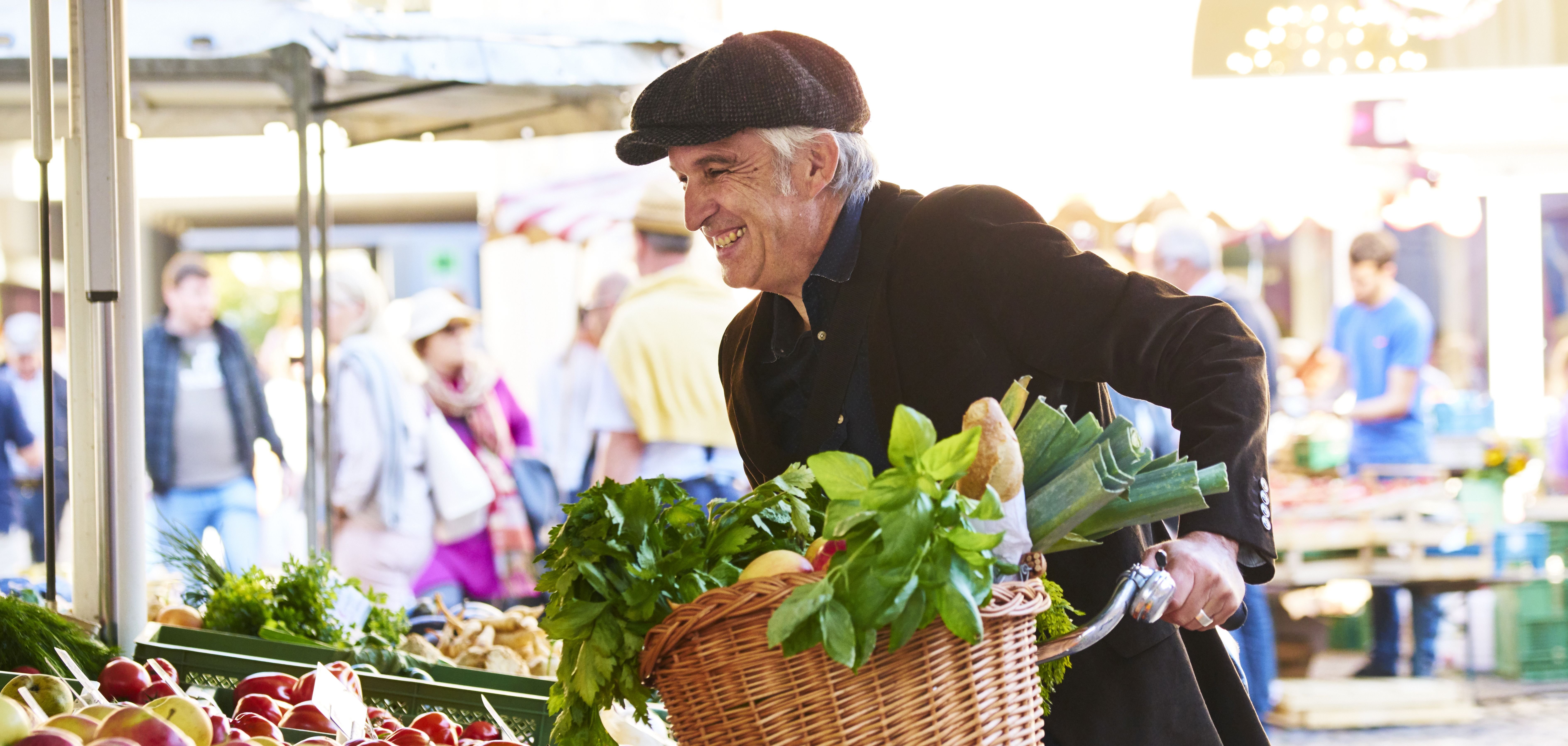 Ein Mann mit Fahrrad auf dem Markt in einer Stadt mit seinen Einkäufen im Fahrradkorb, darunter Baguette, Gemüse, Obst.