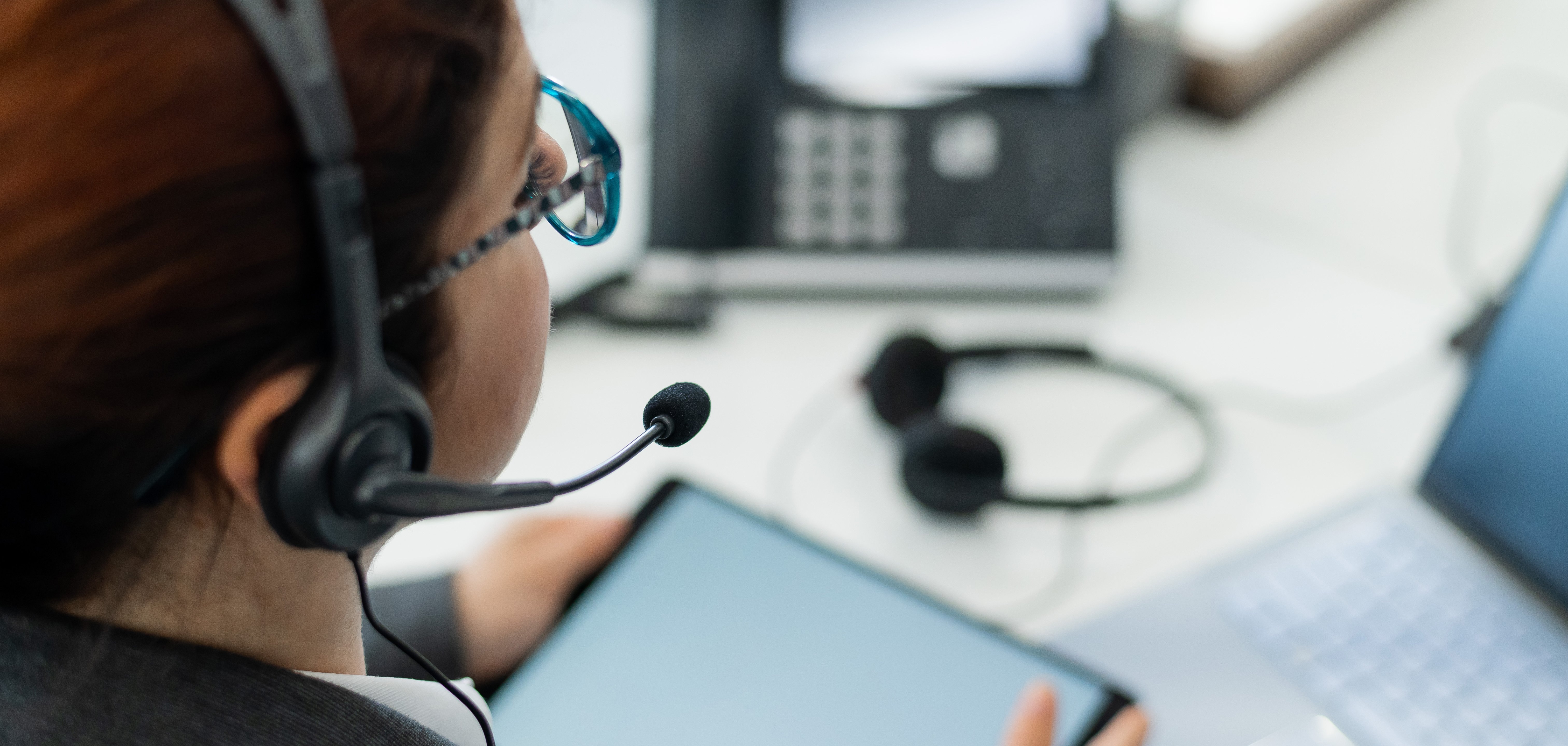 Eine Frau sitzt an einem Bürotisch, hat ein Tablet in der Hand und telefoniert über ein Headset.