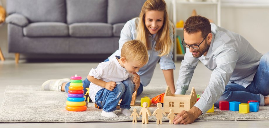 Ein junges Elternpaar sitzt mit einem Kleinkind auf dem Boden ihres Wohnzimmers und spielen mit Bausteinen, beziehungsweise Holzspielzeug.