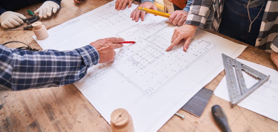 Architekten und Handwerker schauen auf einen Grundriss für ein Hausbau Projekt - zu sehen sind nur ihre Hände, die jeweils die für sie typischen Werkzeuge oder Utensilien halten.
