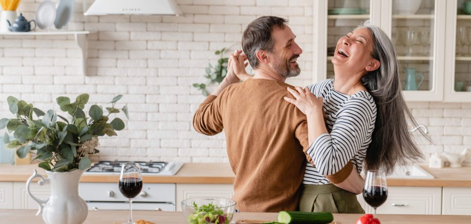 Ein glückliches Paar mittleren Alters tanzt zusammen in der Küche, im Vordergrund sind Kochzutaten für eine gemeinsam zubereitete Mahlzeit zu sehen.