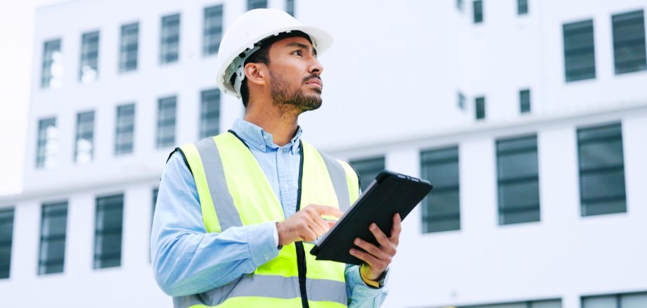 Ein männlicher Architekt oder Ingenieur kontrolliert auf einem Tablet eine Baustelle. Er trägt einen gelben Schutzhelm.