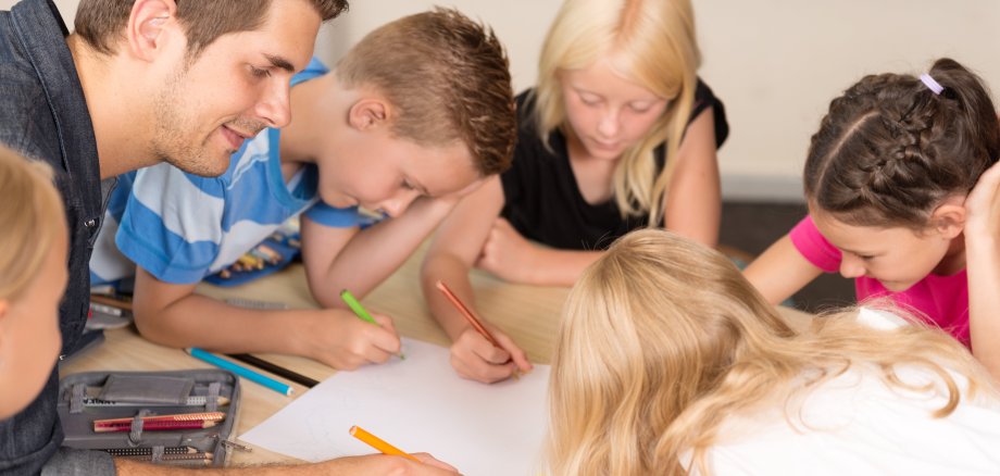 Das Bild zeigt einen Betreuer, der mit mehreren Grundschulkindern bei der Nachmittagsbetreuung am Tisch sitzt und mit ihnen malt.