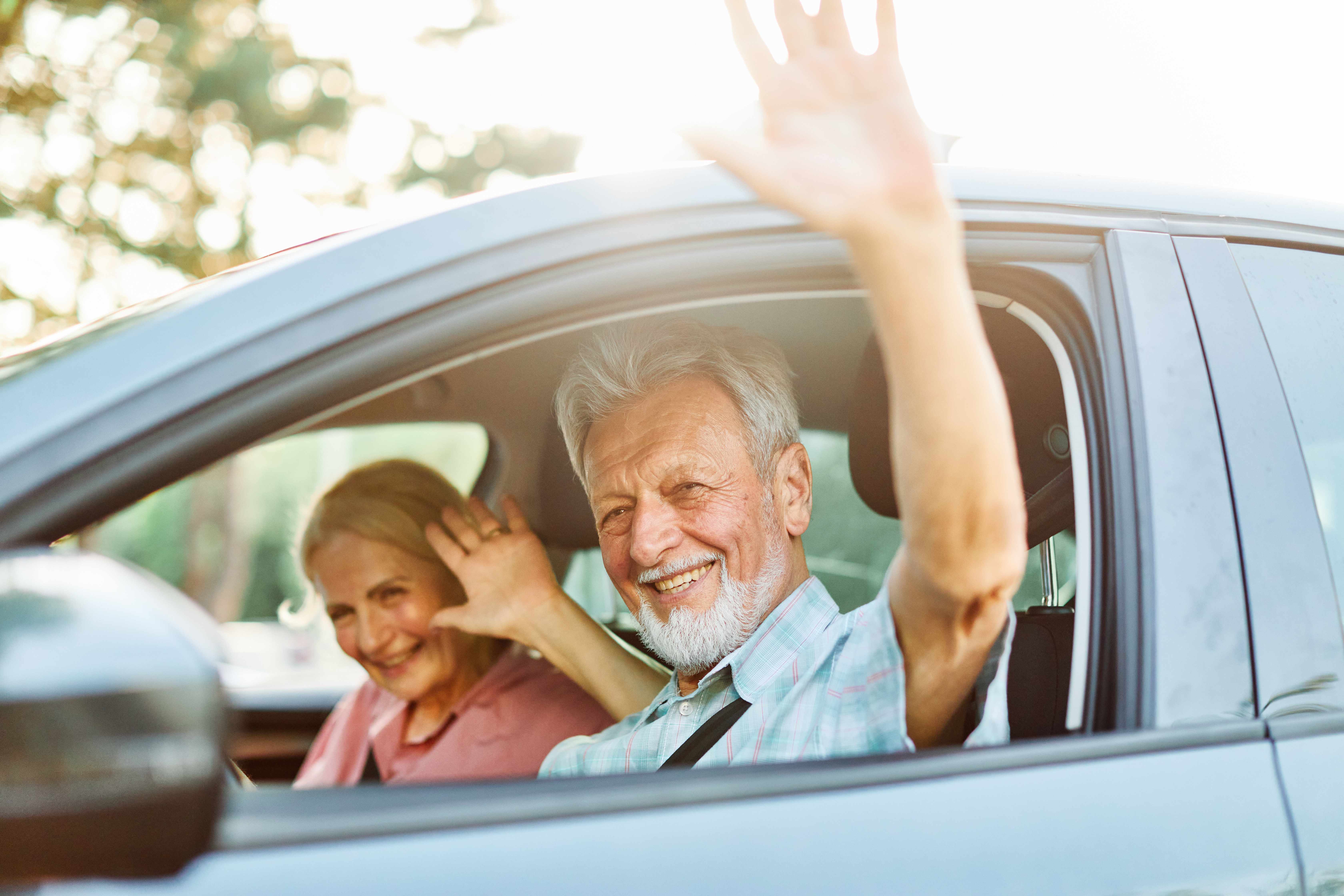 Ein älteres Paar sitzt in seinem Auto und winkt freundlich aus dem Fenster.