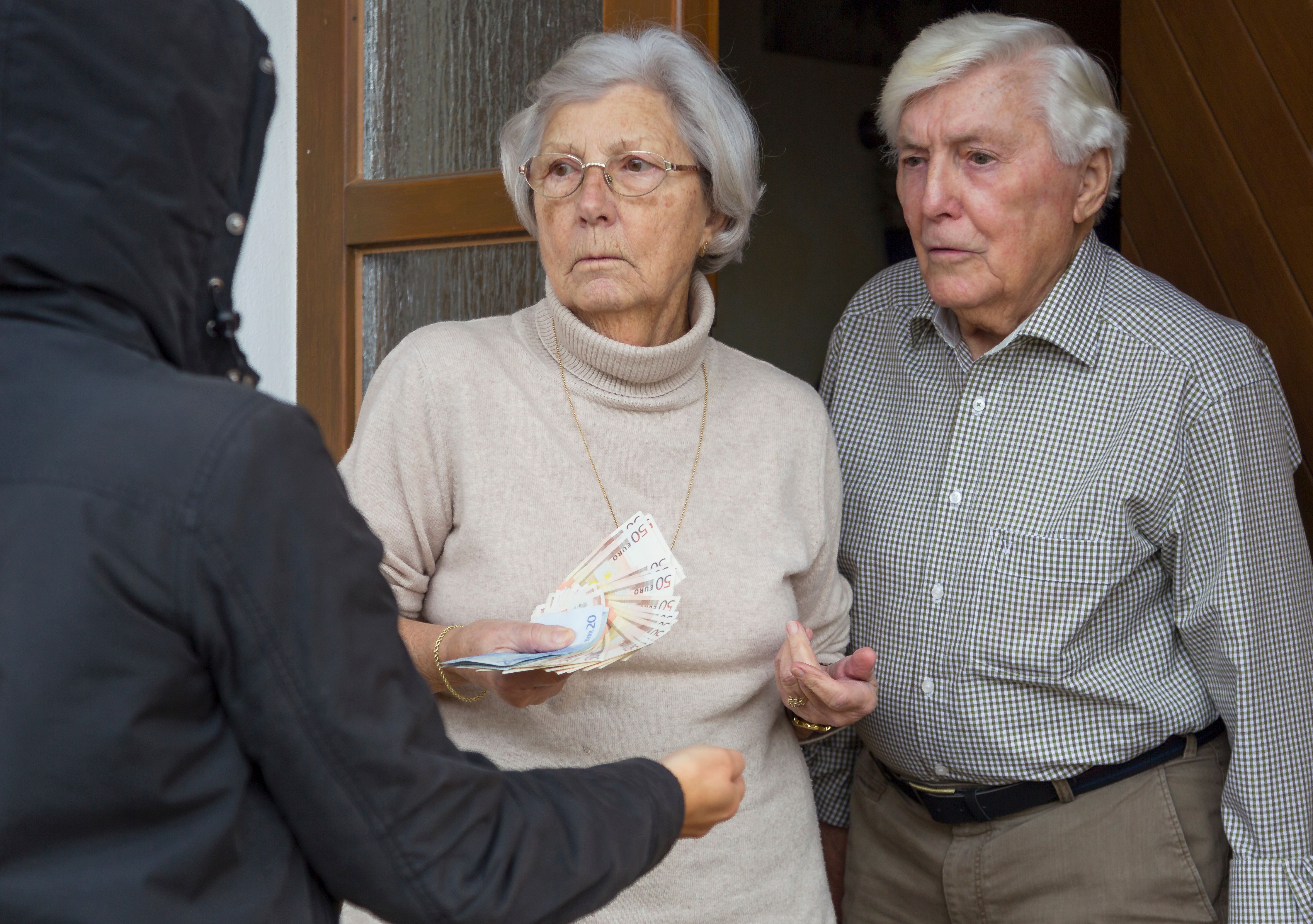 Ein Seniorenpaar steht mit Bargeld in der Hand in der Haustür. Vor ihnen steht eine Person mit Kapuzenpullover, die fordern die Hand ausstreckt.