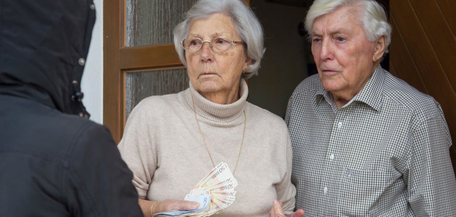 Ein Seniorenpaar steht mit Bargeld in der Hand in der Haustür. Vor ihnen steht eine Person mit Kapuzenpullover, die fordern die Hand ausstreckt.