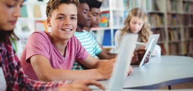 Fünf Schülerinnen und Schüler sitzen in einer Bibliothek und arbeiten an Laptops. Ein Junge in einem roten T-Shirt lächelt in die Kamera.