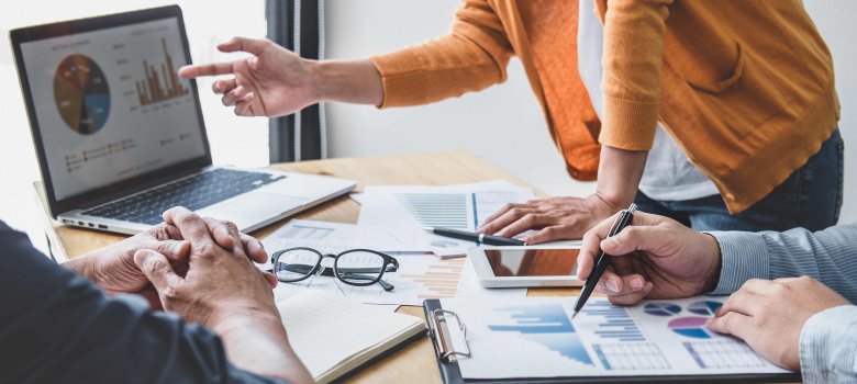 Arbeitsgruppe im Büro Ein Tisch auf dem ein Laptop steht, verschiedene Arbeitsunterlagen sowie eine Brille liegen. Drei Personen sind um den Tisch platziert, man sieht jedoch nur ihre Arme und Hände, wie diese auf die Unterlagen oder die Graphen auf dem Computerbildschirm deuten.