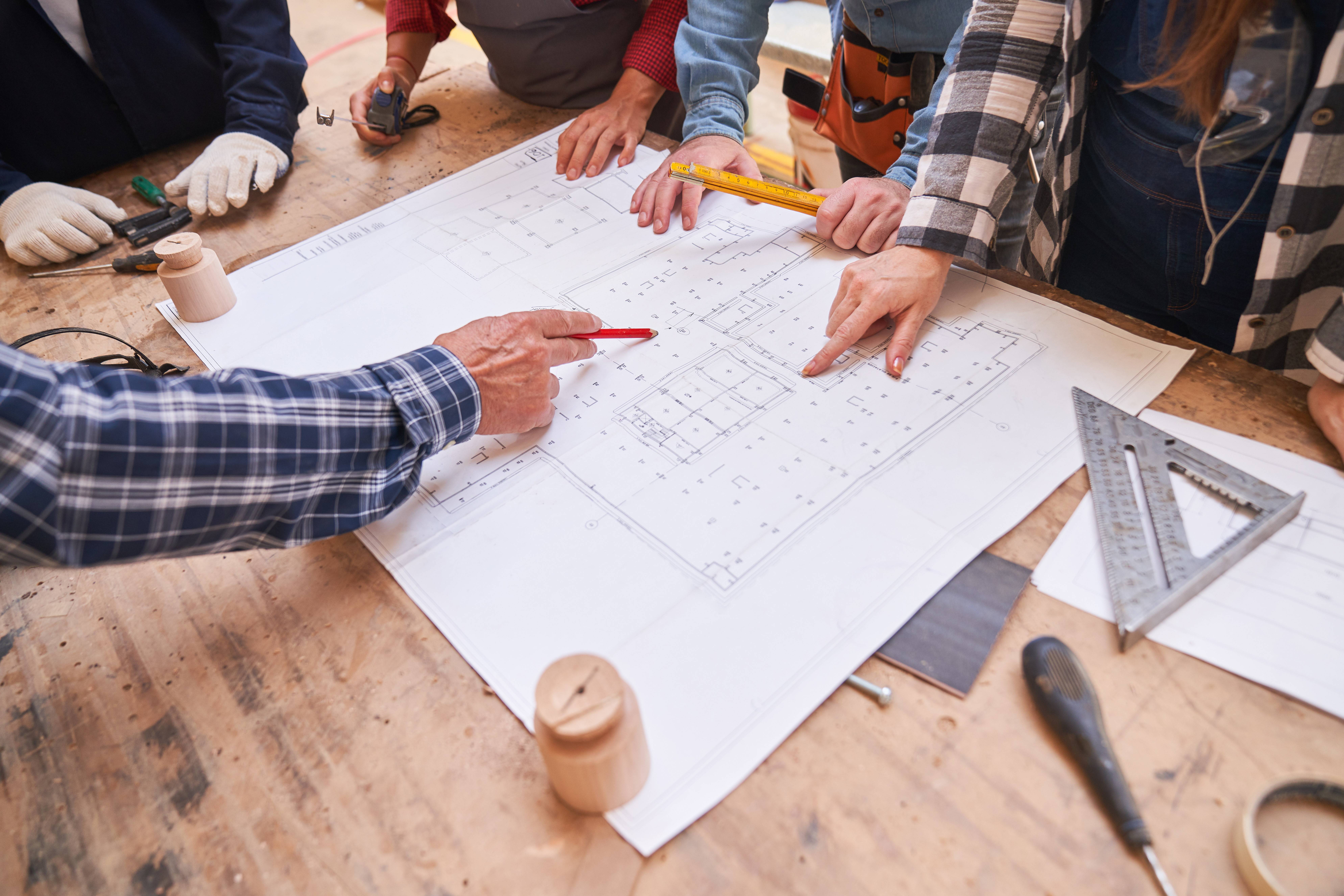 Architekten und Handwerker schauen auf einen Grundriss für ein Hausbau Projekt - zu sehen sind nur ihre Hände, die jeweils die für sie typischen Werkzeuge oder Utensilien halten.