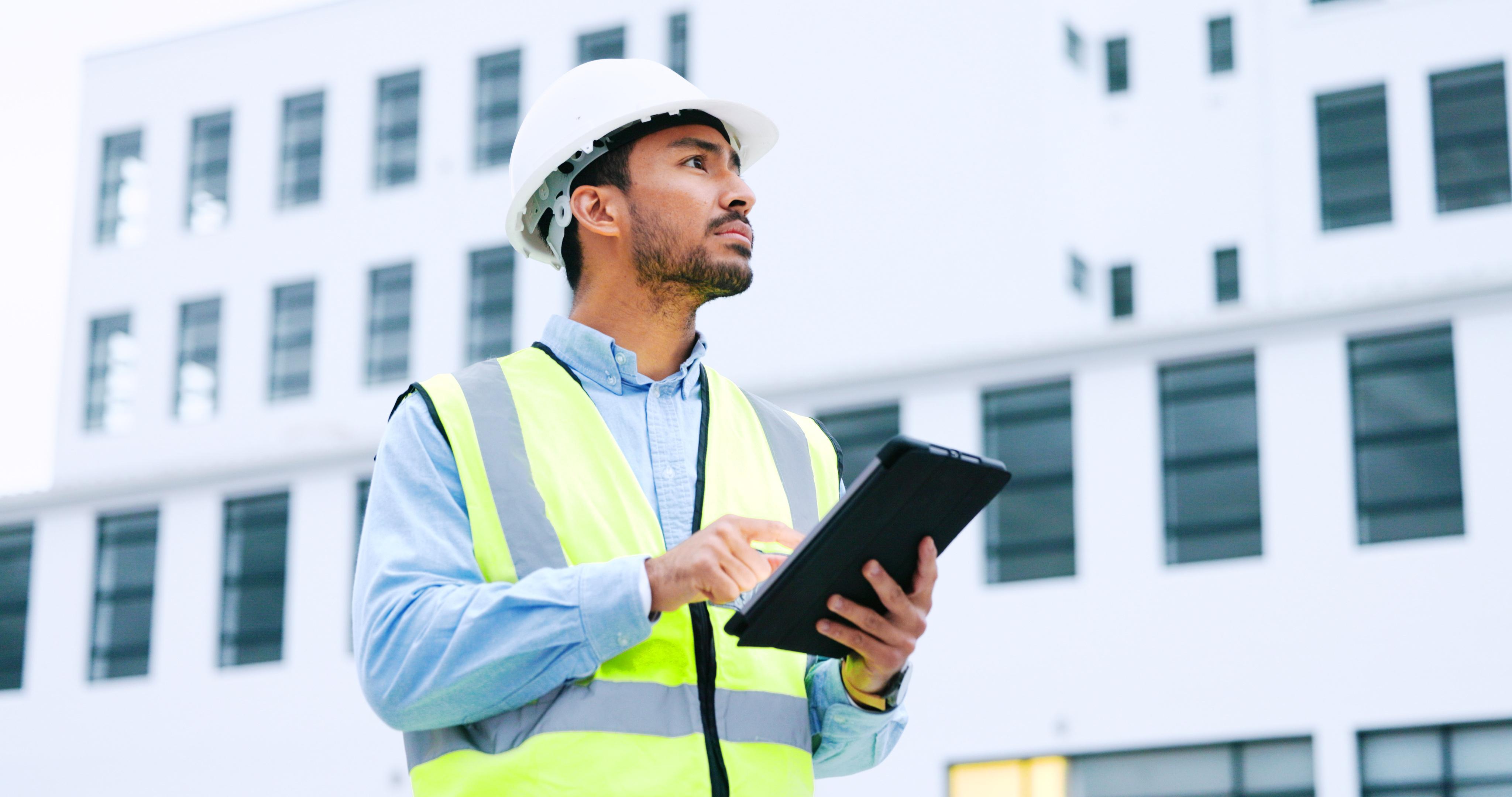 Ein männlicher Architekt oder Ingenieur kontrolliert auf einem Tablet eine Baustelle. Er trägt einen gelben Schutzhelm.