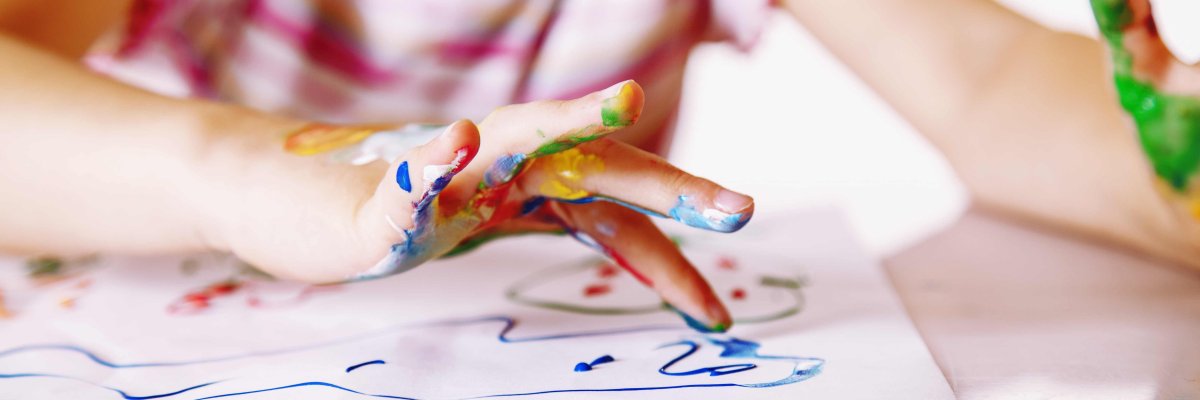 Nahaufnahme von einer Kinderhand, die mit Fingerfarben etwas auf ein weißes Blatt Papier malt.