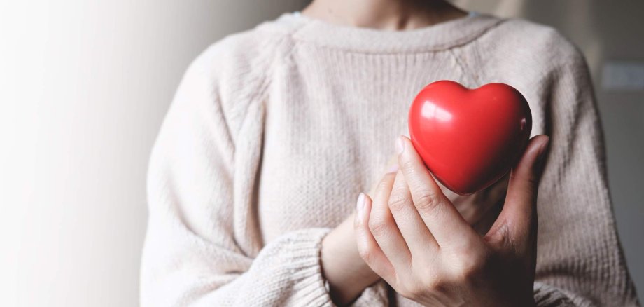 Eine Frau hält auf Höhe des Herzens einen roten Stein, in der Form eines Herzens.