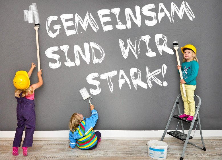 drei Kinder malen mit weißer Farbe den Schriftzug "gemeinsam sind wir stark" auf eine graue Wand.