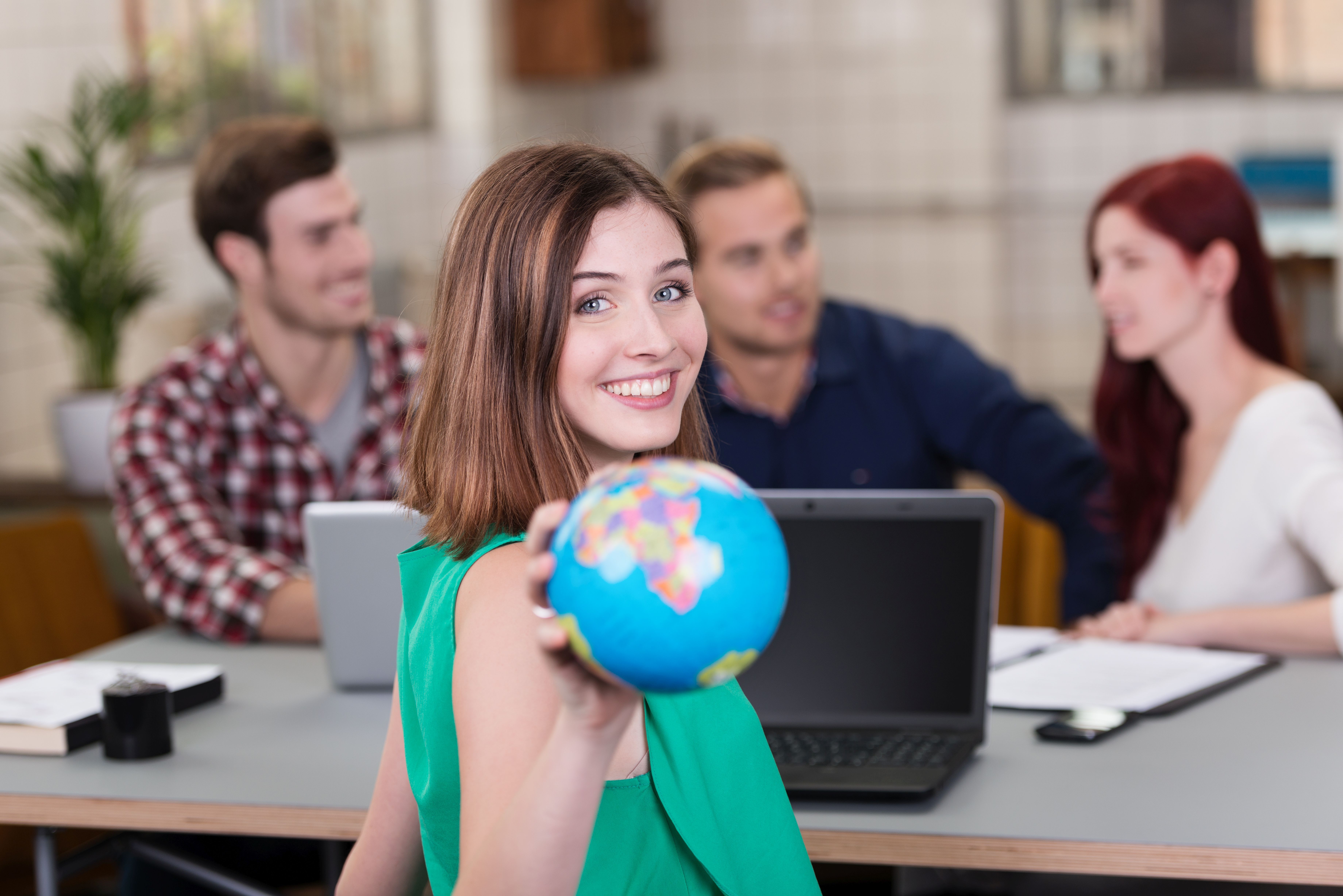 junge frau zeigt weltkugel EIne junge Frau hält eine Weltkigel in der Hand. Im Hintergrund arbeiten drei junge Menschen an Laptops
