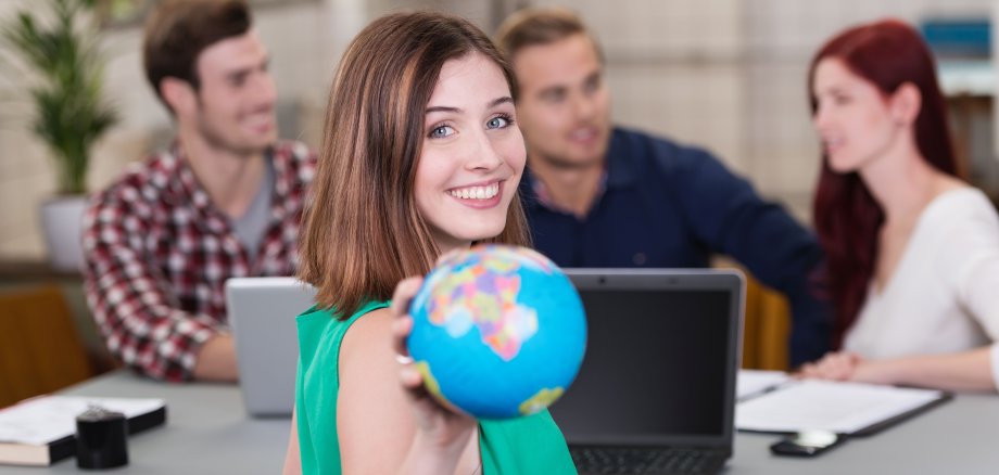 EIne junge Frau hält eine Weltkigel in der Hand. Im Hintergrund arbeiten drei junge Menschen an Laptops 