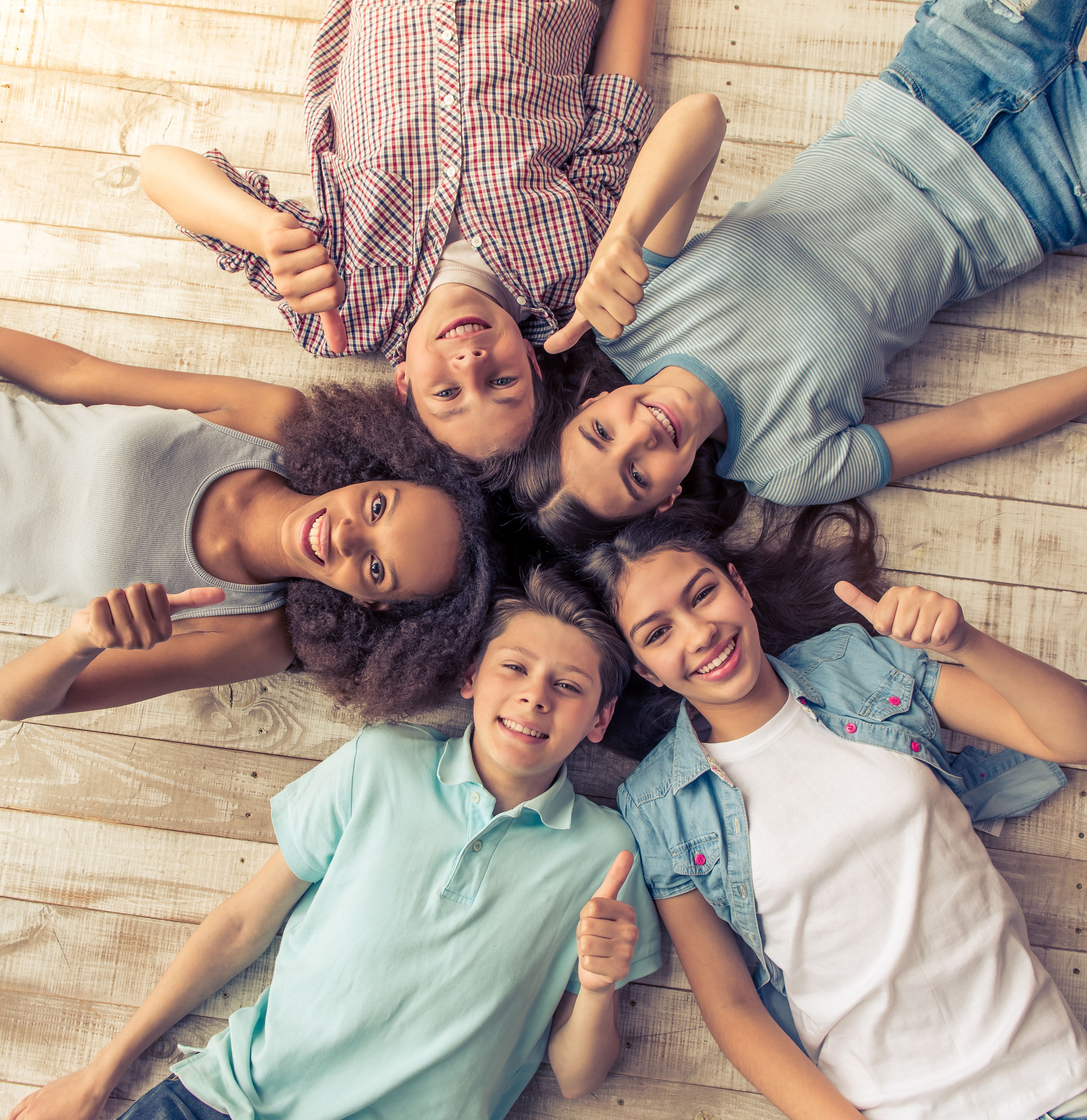 Jugendliche liegen in einem Kreis, die Köpfe berühren sich in der Mitte. Alle haben eine Hand mit dem Daumen nach oben erhoben.
