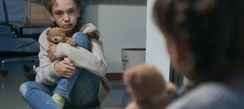 Mädchen mit Verletzungen im Gesicht sitzt vor einem Spiegel und umarmt Plüschbär.