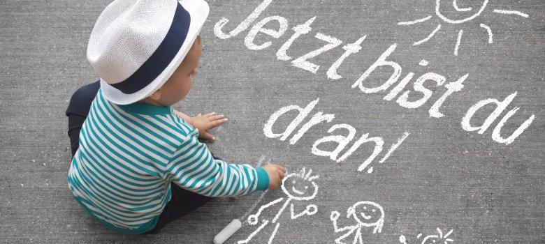 Kinderzeichnung - Jetzt bist du dran! Kleinkind schreibt mit Kreide auf den Boden "Jetzt bist du dran!"