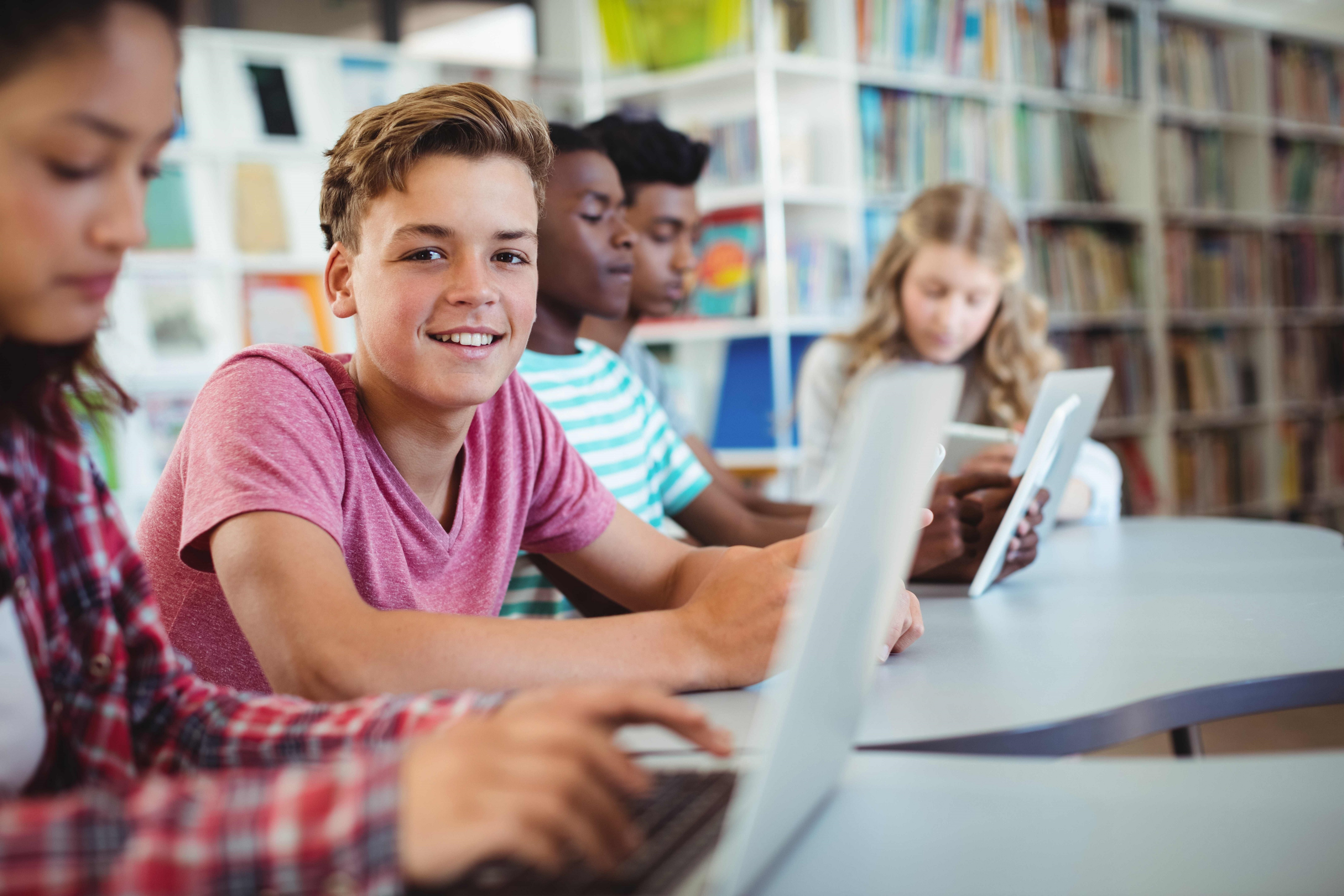 Arbeiten mit Schullaptops Fünf Schülerinnen und Schüler sitzen in einer Bibliothek und arbeiten an Laptops. Ein Junge in einem roten T-Shirt lächelt in die Kamera.