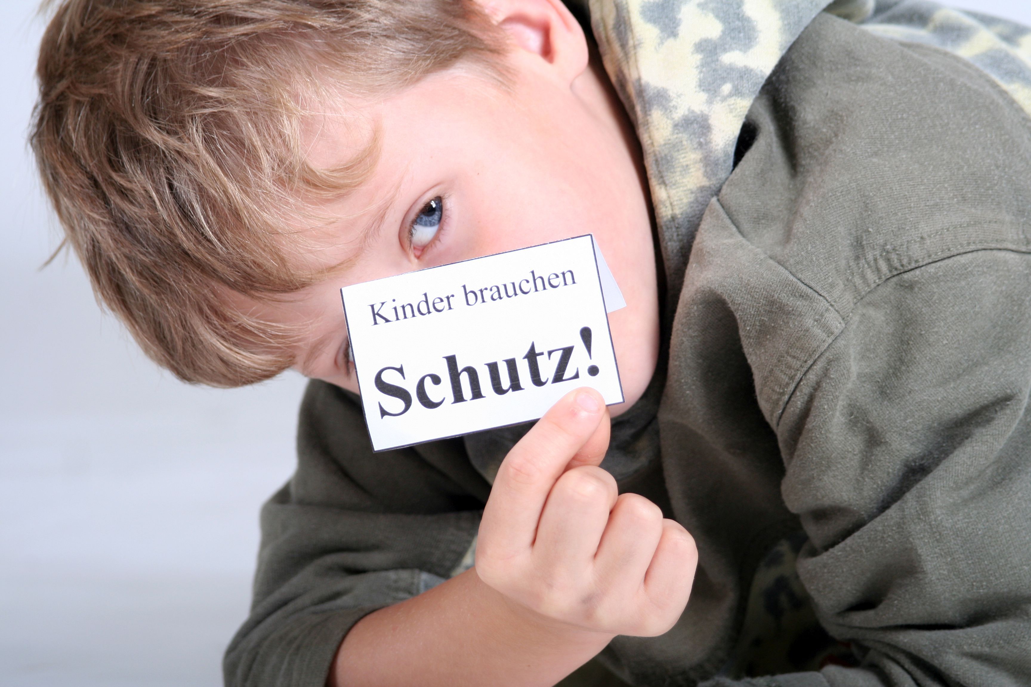 Ein Junge hält ein kleines weißes Stück Papier mit der Aufschrift "Kinder brauchen Schutz!" vor sein Gesicht. 