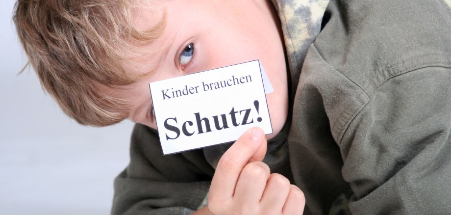 Kinderschutz Ein Junge hält ein kleines weißes Stück Papier mit der Aufschrift "Kinder brauchen Schutz!" vor sein Gesicht.