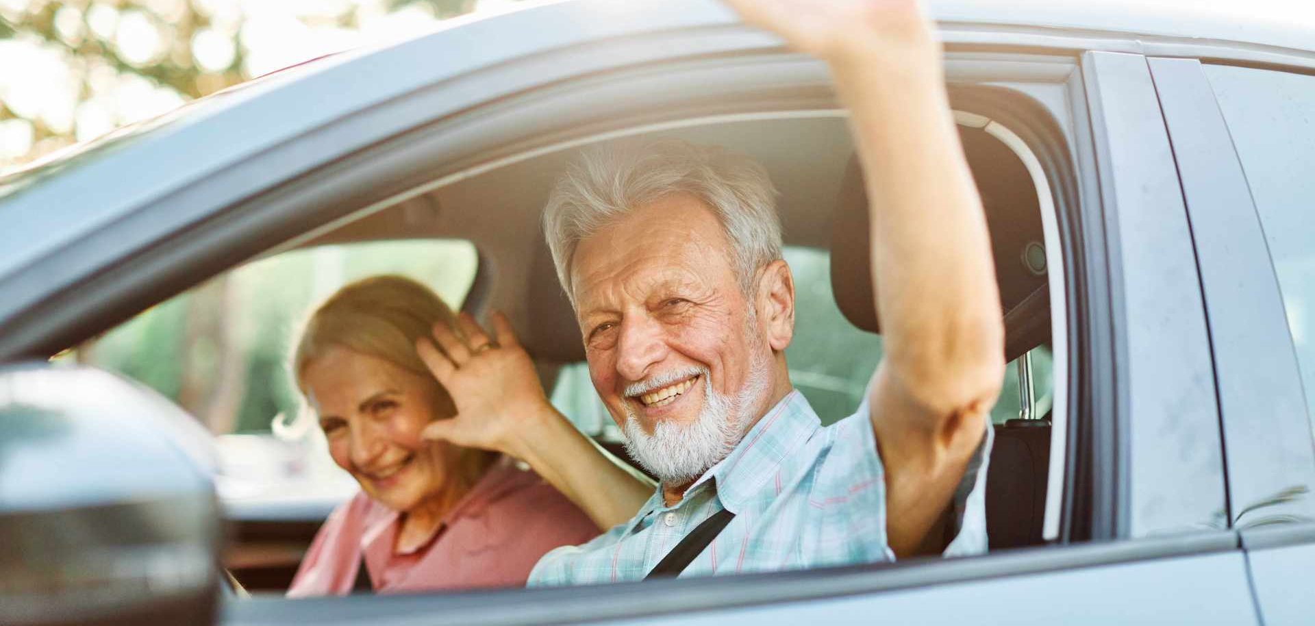 Ein lächelndes Seniorenpaar sitzt in einem Auto und winkt aus dem Fenster.