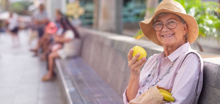 Eine Seniorin sitzt auf einer Bank in der Sonne mit einem Apfel in der Hand.