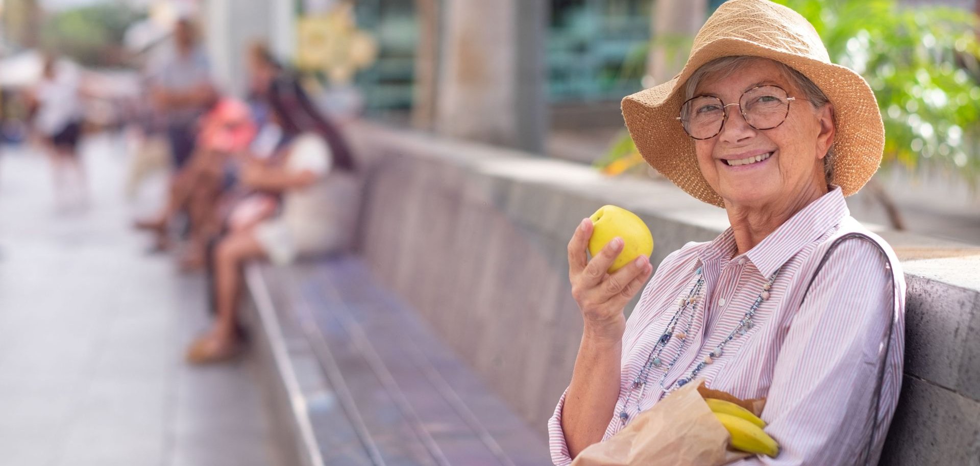 Eine Seniorin sitzt auf einer Bank in der Sonne mit einem Apfel in der Hand.