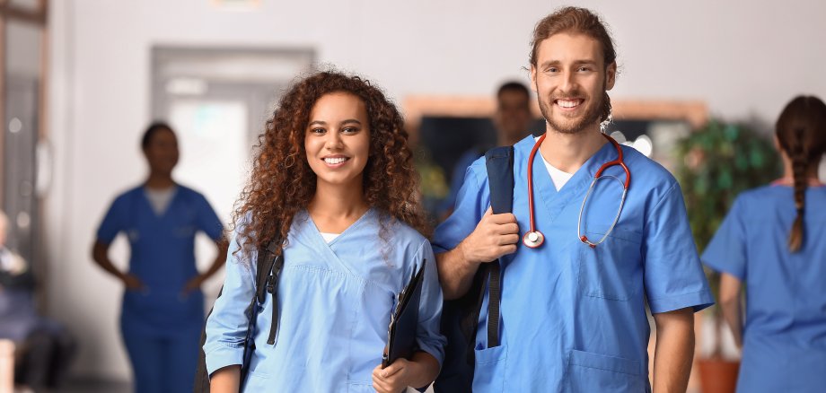 Zwei Medizinstudierende stehen in einem Flur und lächeln in die Kamera.