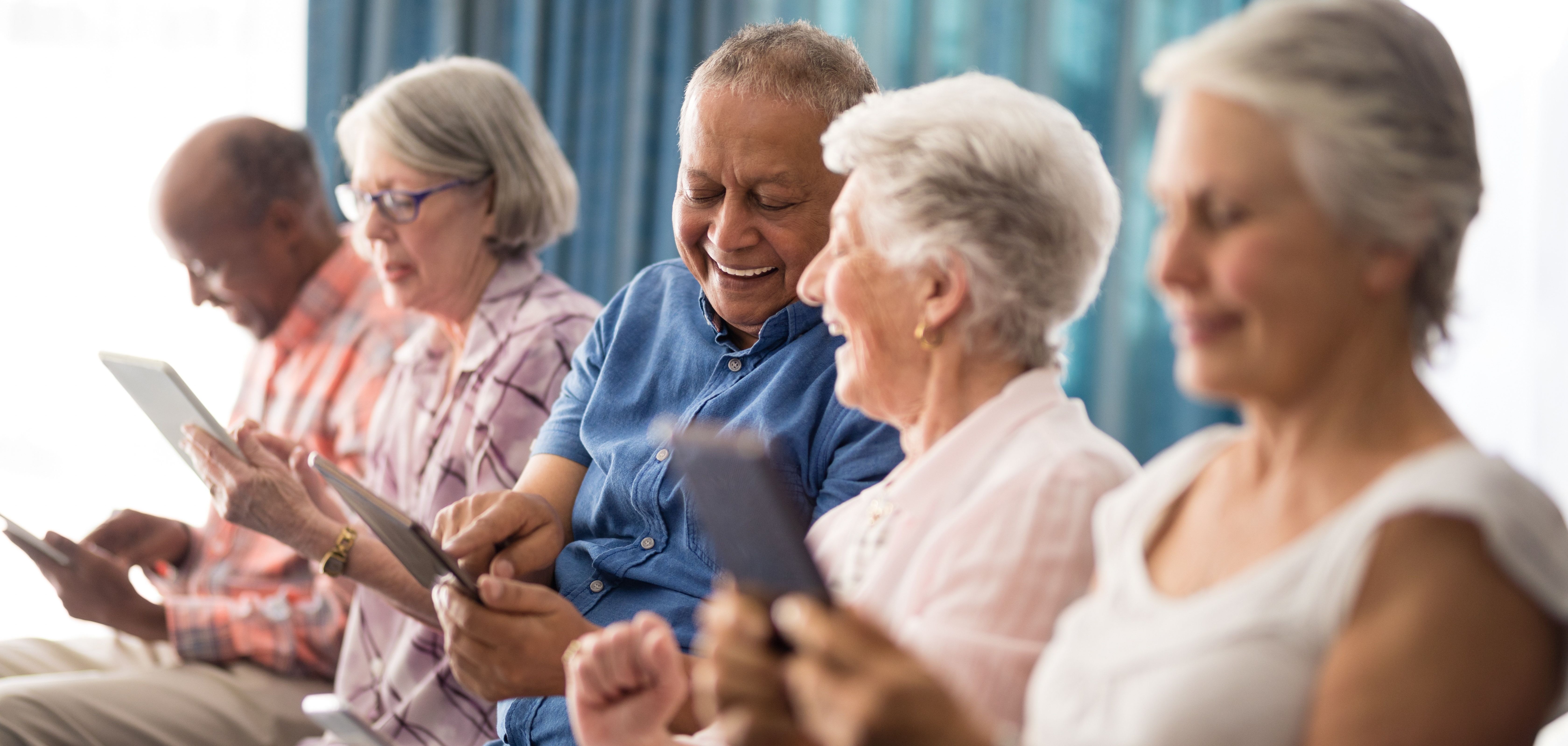 Lächelnde Seniorinnen und Senioren sitzen in einer Reihe und halten Handys und Tablets in den Händen.