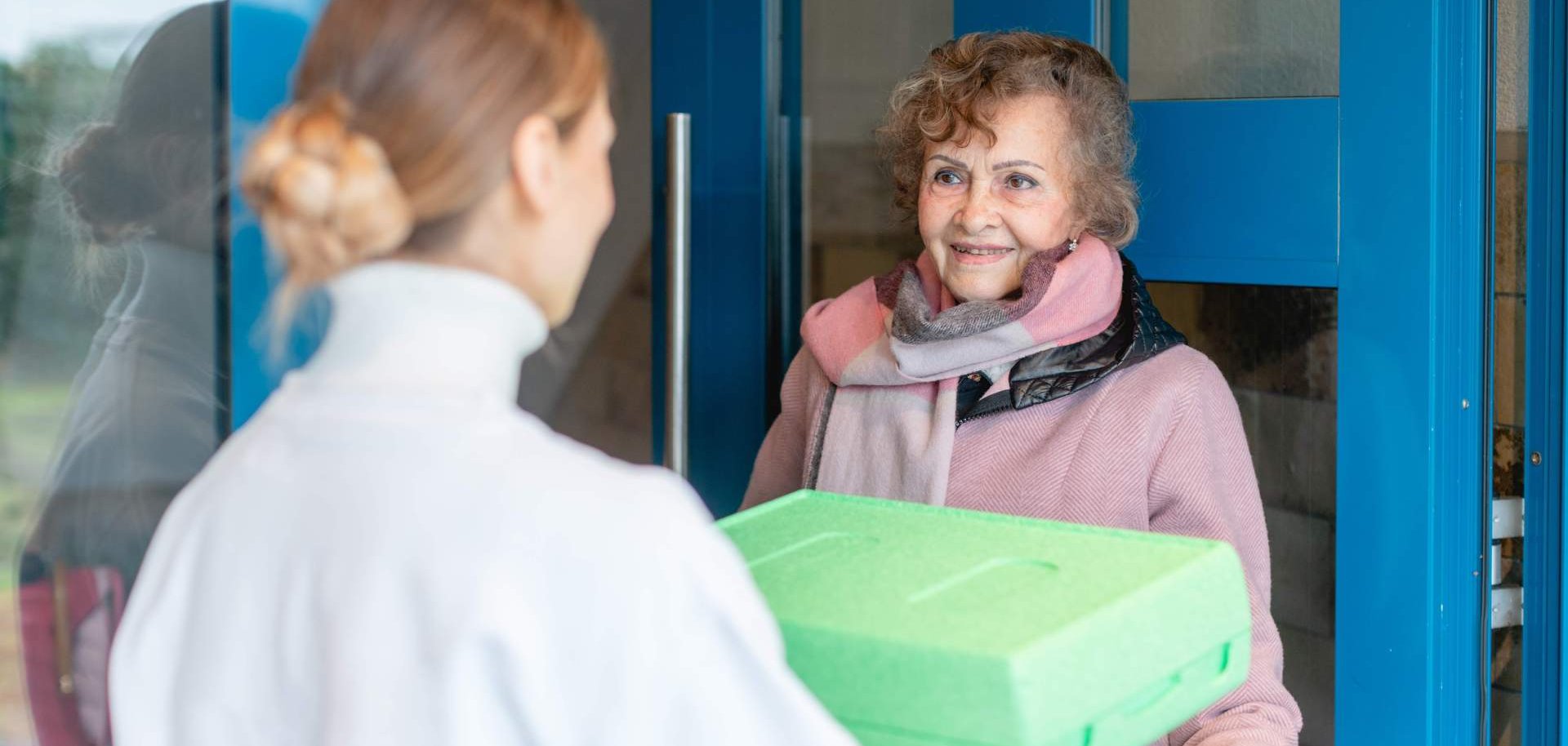 Eine Frau übergibt einer Seniorin eine verpackte Mahlzeit an ihrer Haustür.