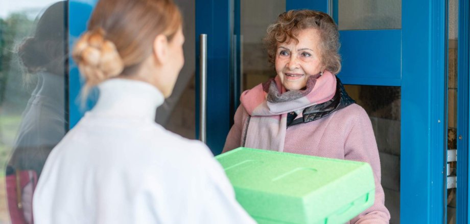 Eine Frau übergibt einer Seniorin eine verpackte Mahlzeit an ihrer Haustür.
