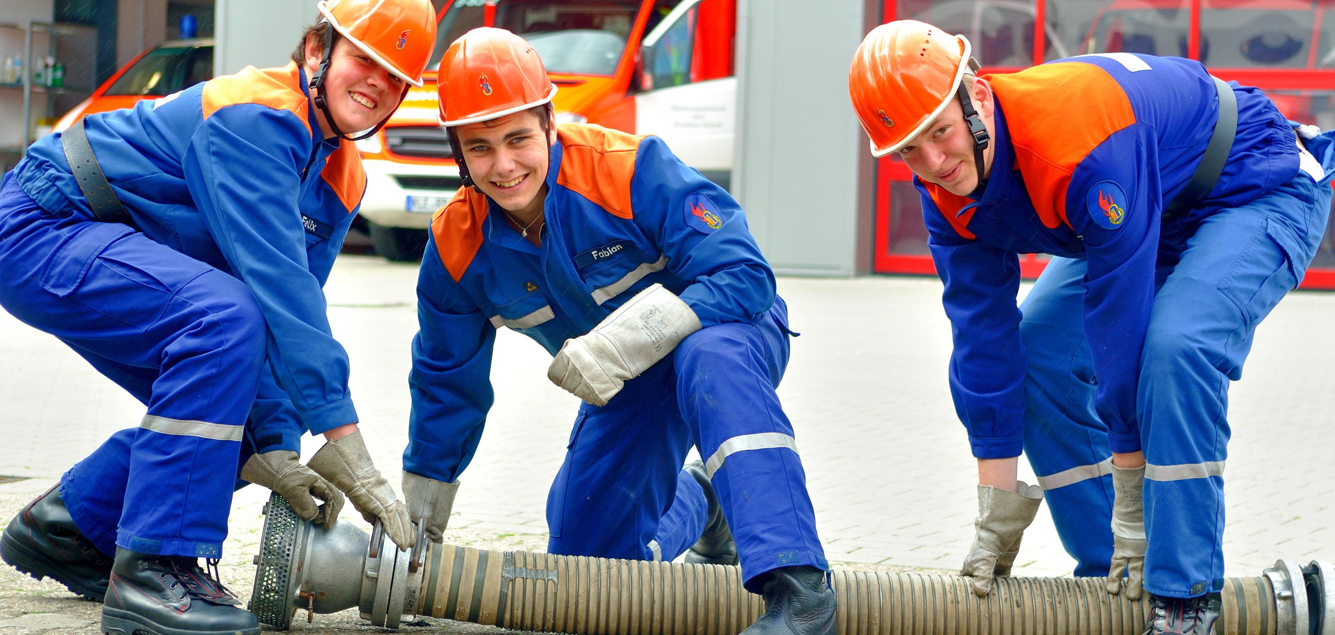 Drei junge Männer der Freiwilligen Feuerwehr knien vor einem Feuerwehrhaus um einen Wasserschlauch und lachen in die Kamera.