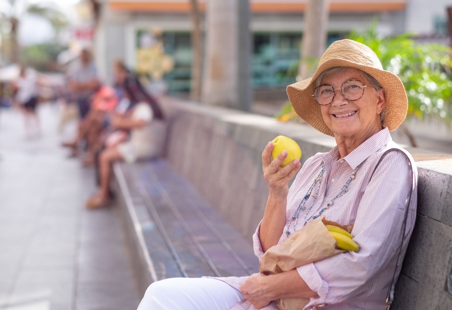 Eine Seniorin sitzt lächelnd mit Einkäufen auf einer Parkbank und hält einen Apfel in der Hand.