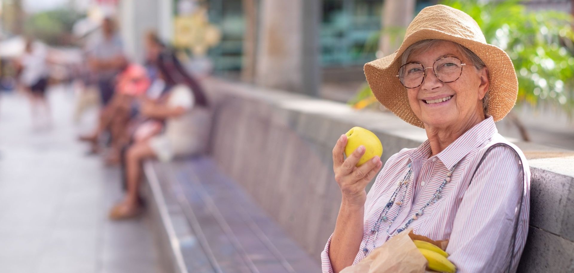 Eine Seniorin sitzt lächelnd mit Einkäufen auf einer Parkbank und hält einen Apfel in der Hand.
