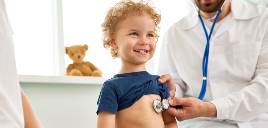 Ein kleiner, lächelnder Junge wird von einem Arzt mit einem Stethoskop abgehört.