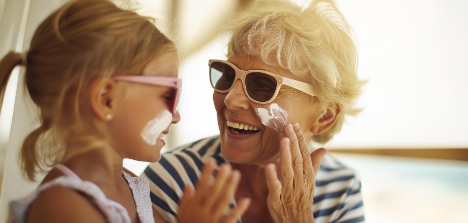 Grandmother applying sunscreen to her granddaughter at the beach. Summer vacation, skin protection, sun care, and loving family moment under bright sunlight by the ocean.