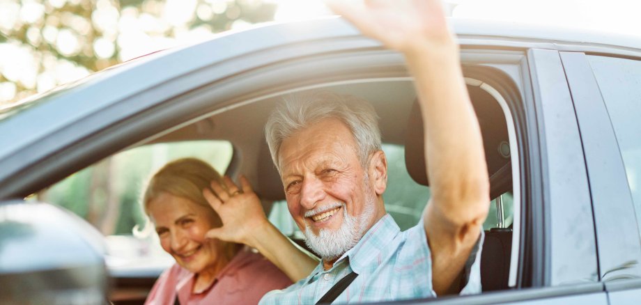 Ein älteres Paar sitzen gemeinsam in einem Auto und winken lächelnd.