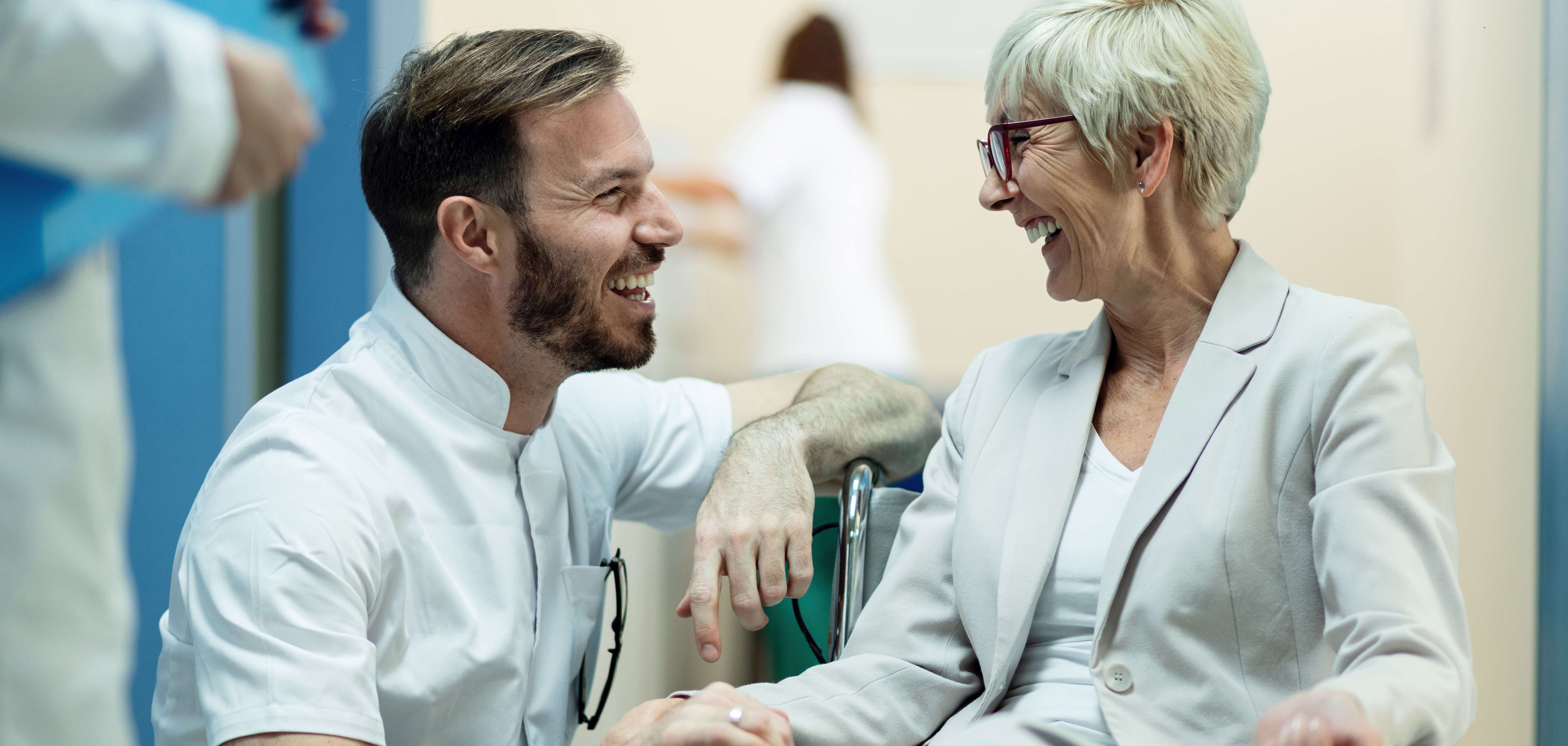 Eine fröhliche Seniorin sitzt in einem Rollstuhl und unterhält sich lachend mit einem Pfleger, der sich zu ihr runter gehockt hat.