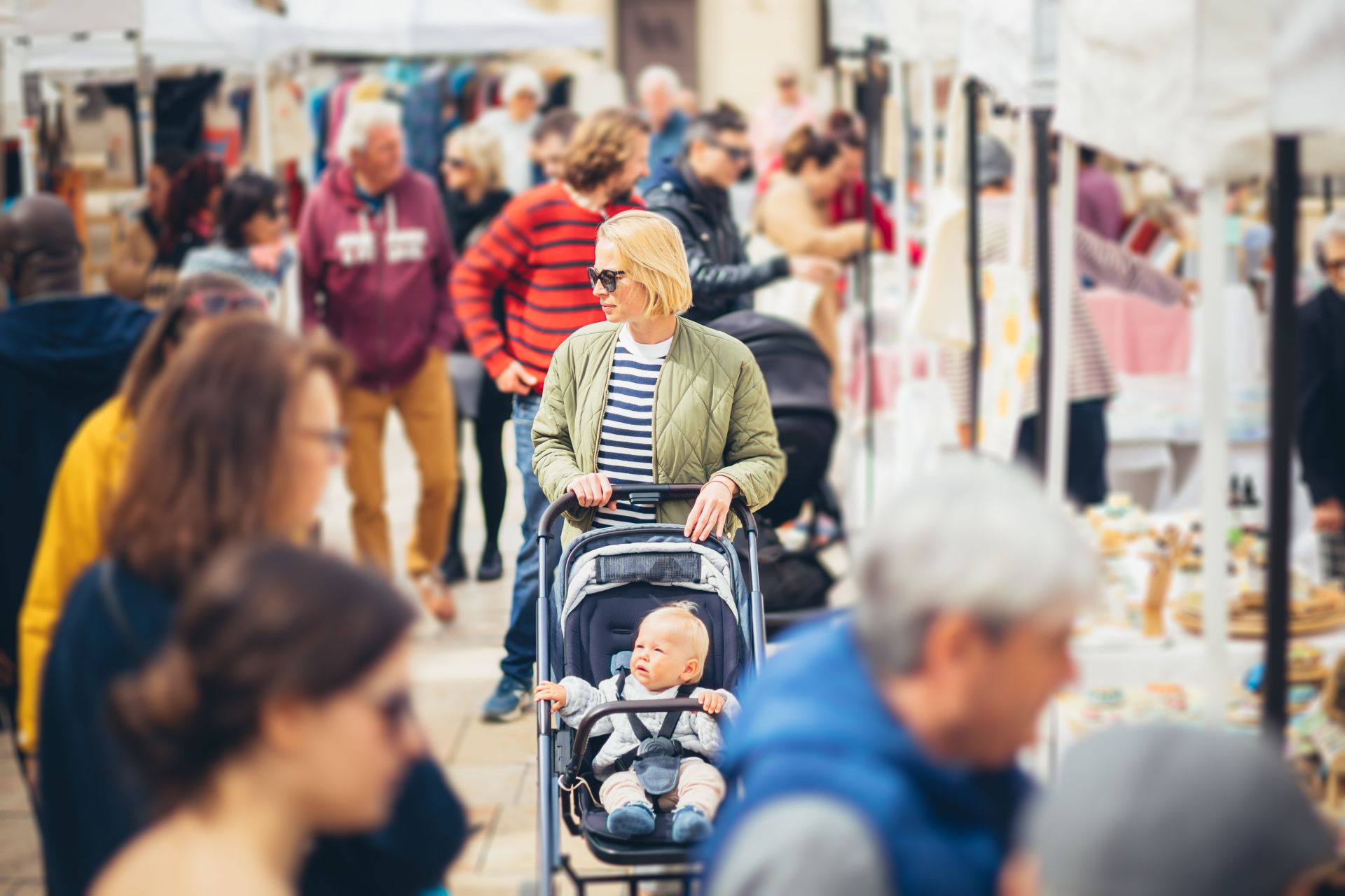 Mother waling and pushing his infant baby boy child in stroller in crowd of unrecognizable people wisiting sunday flea market in Malaga, Spain.
