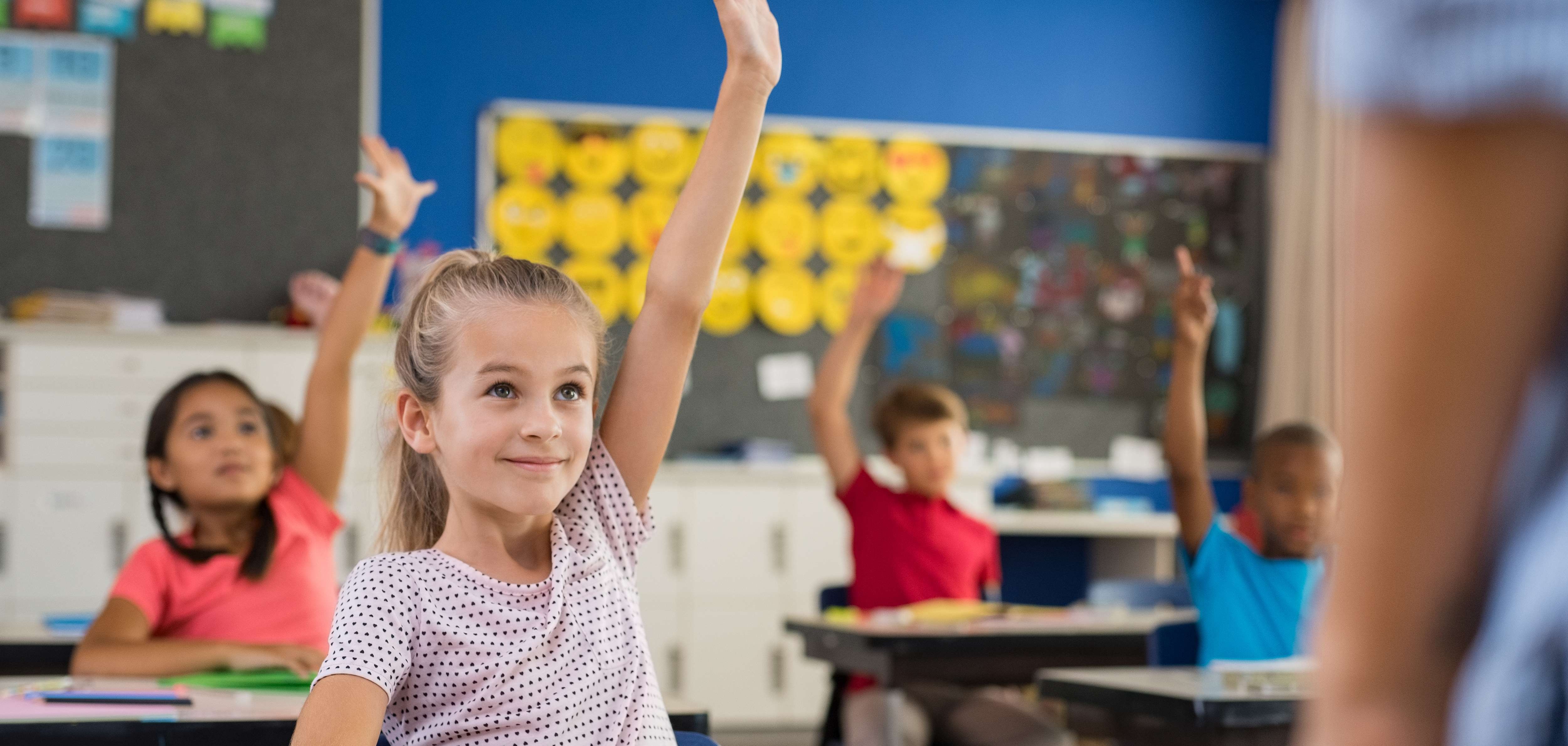 Mehrere Grundschulkinder sitzen in einem Klassenraum. Ein Mädchen meldet sich eifrig.