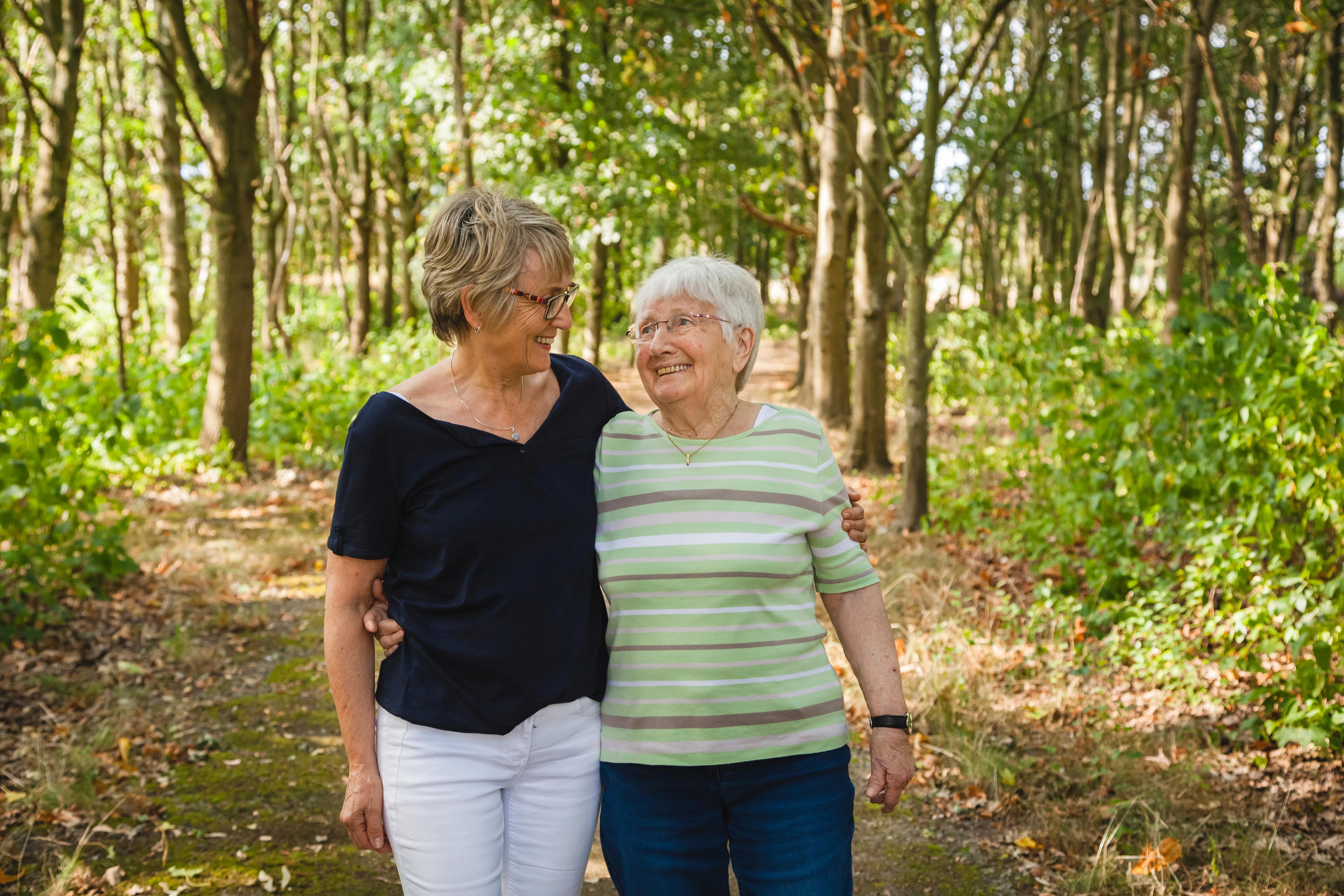 Eine fröhliche Seniorin geht mit einer jüngeren Frau im Wald spazieren. Die Jüngere hat den Arm um die Ältere gelegt.