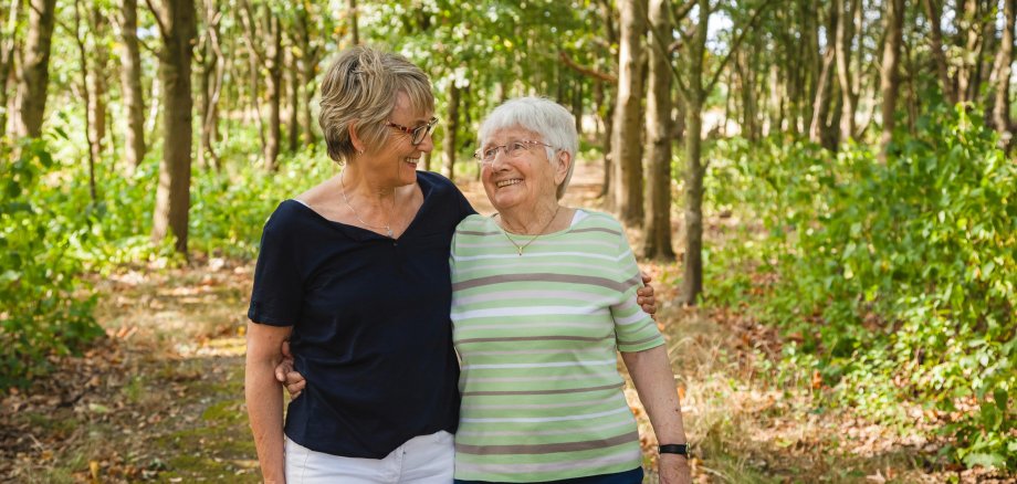 Eine fröhliche Seniorin geht mit einer jüngeren Frau im Wald spazieren. Die Jüngere hat den Arm um die Ältere gelegt.