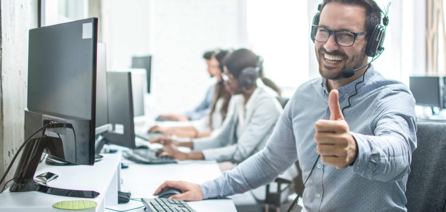 Ein dunkelhaariger Mann mit Vollbart, Brille und Headseit sitzt am Computer, lächelt in die Kamera und hält den Daumen hoch.