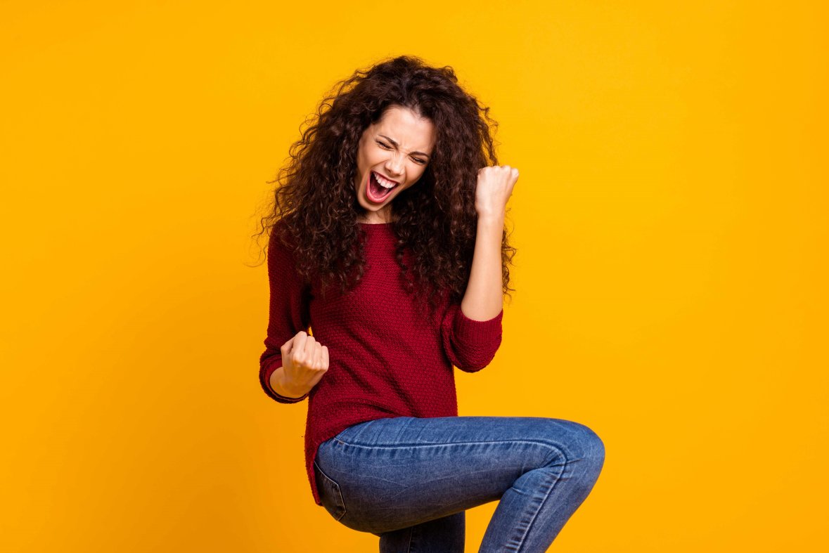 Jungefrau mit dunklen Locken, in einem Roten Pullover und blauen Jeans, freut sich vor einem gelben hintergrund.