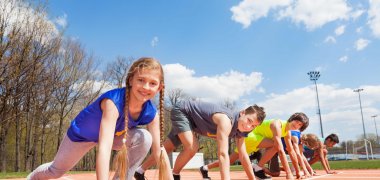 Fünf Kinder sind in Startposition auf einer orangenen Laufbahn unter blauem Himmel.