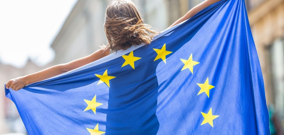 Ein junges Mädchen hält eine ausgebreitete Europaflagge hinter ihrem Rücken.