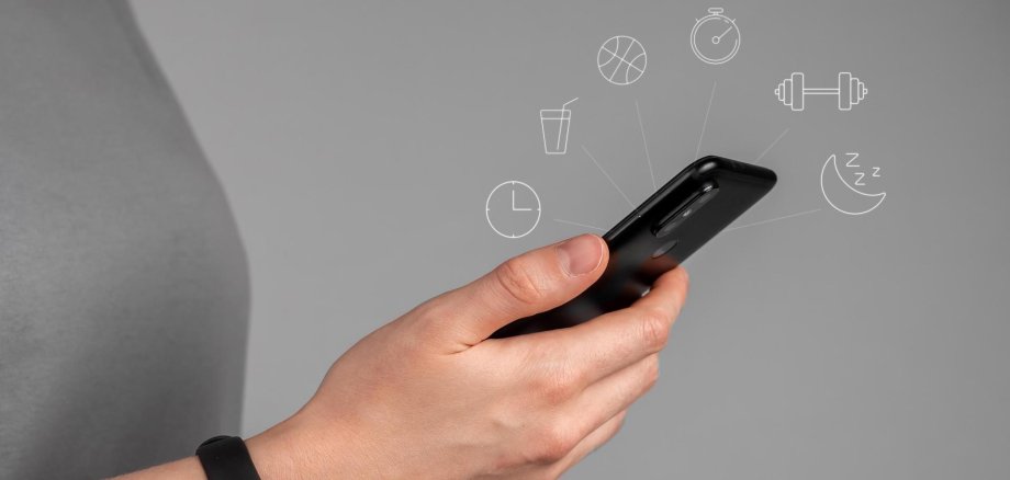 Grenzenlos gesund am 21.10. Eine Hand mit einem Fitnessband hält ein Handy in der Hand, aus dem verschiedene Icons rauszeigen.